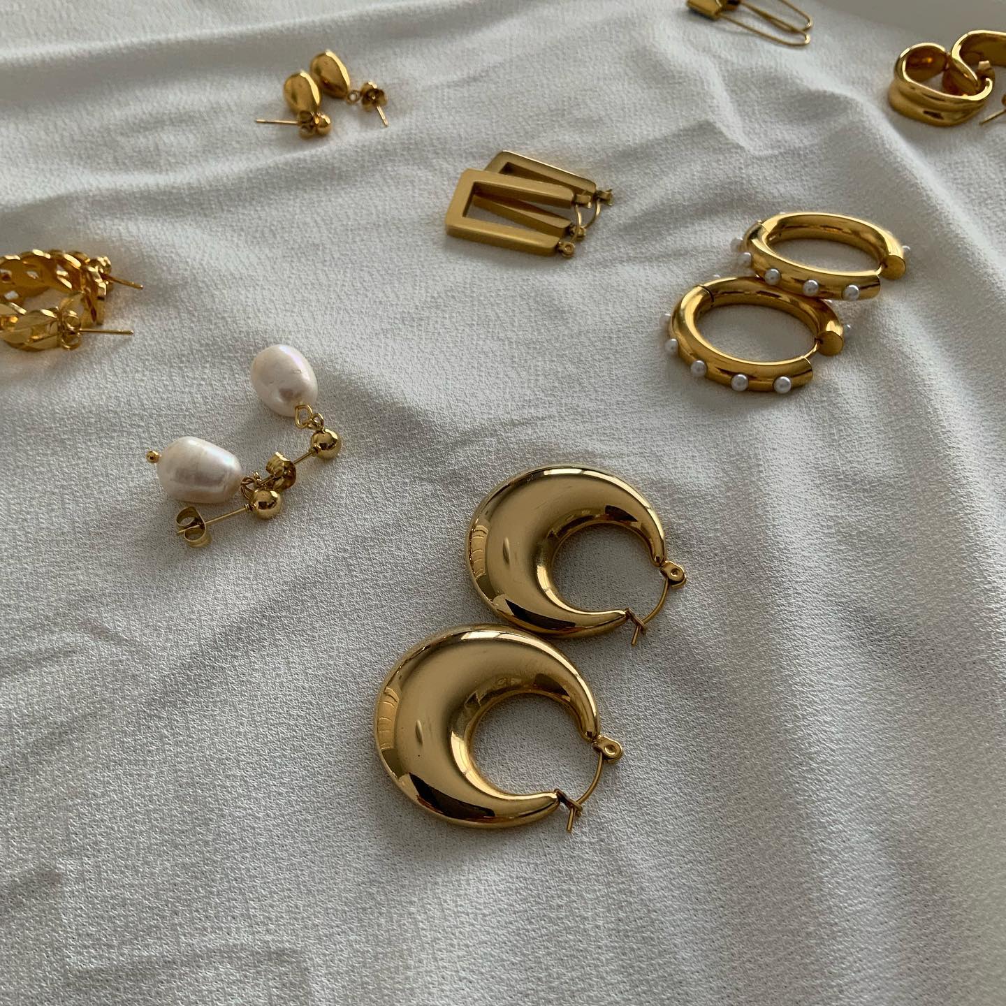 A flat lay of gold hoops, pearl earrings, and charms on cream linen
